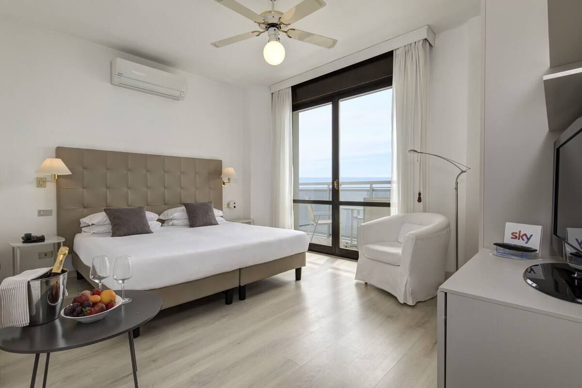 A hotel room with a large bed, a desk with a fruit bowl, a chair, a TV, and a balcony with a view.