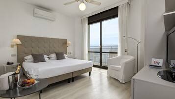 A hotel room with a large bed, a desk with a fruit bowl, a chair, a TV, and a balcony with a view.