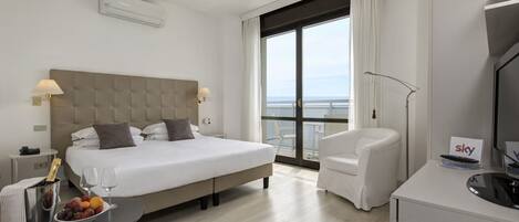 A hotel room with a large bed, a desk with a fruit bowl, a chair, a TV, and a balcony with a view.