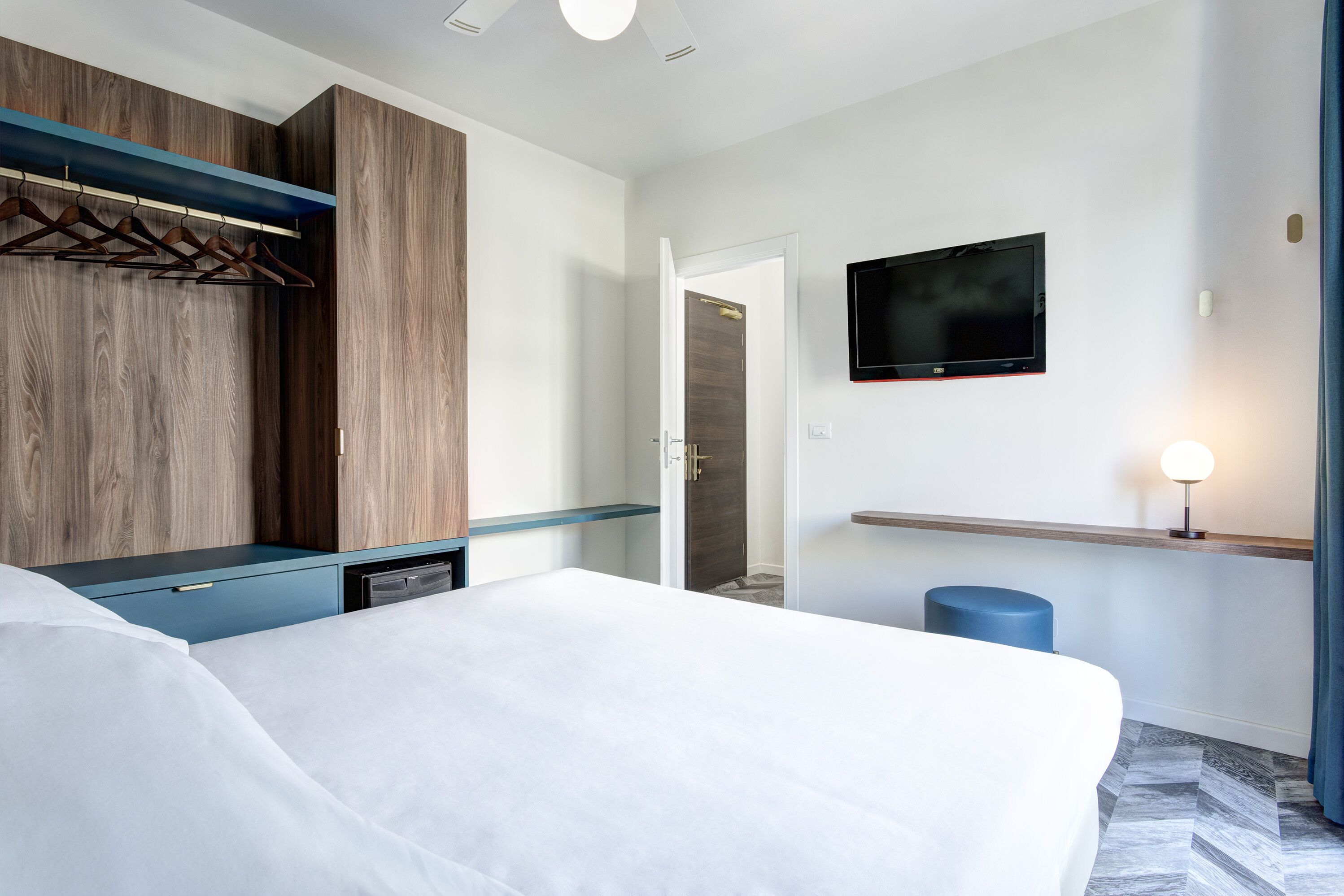 A modern hotel room with a bed, a TV mounted on the wall, a wooden wardrobe, and a blue accent wall.