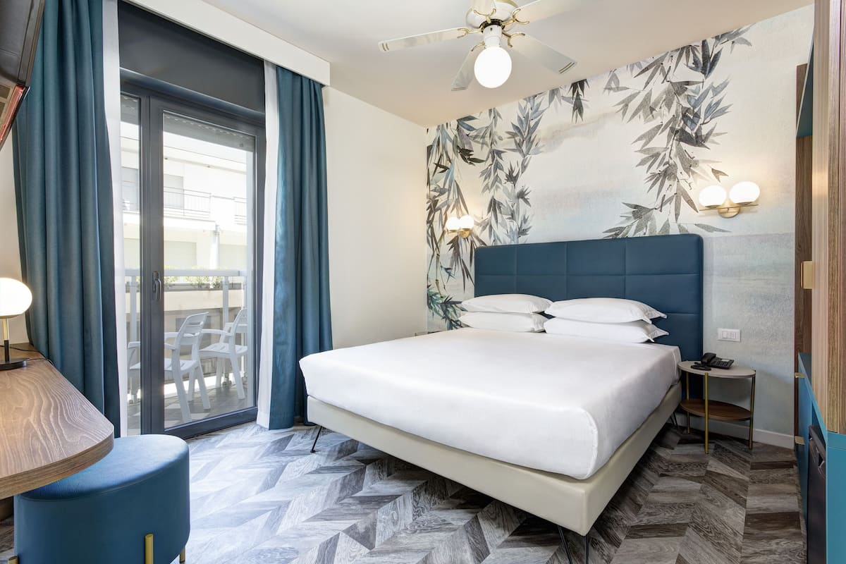 A modern hotel room with a large bed, a ceiling fan, a view through a sliding glass door, and a wall with a bamboo leaf wallpaper.