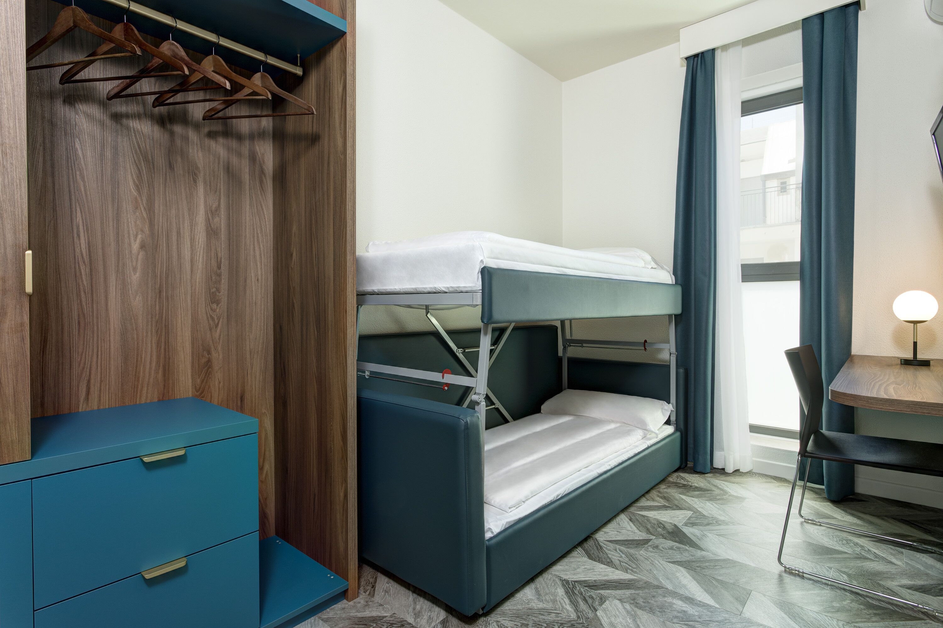A bunk bed with a desk and chair, a wardrobe with hanging clothes, and a window with curtains.