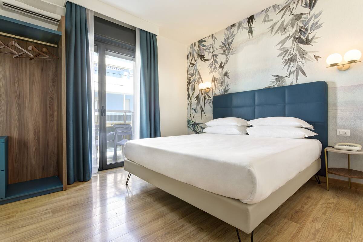 A modern hotel room with a large bed, a wardrobe, a bedside table, and a view of the outside balcony.