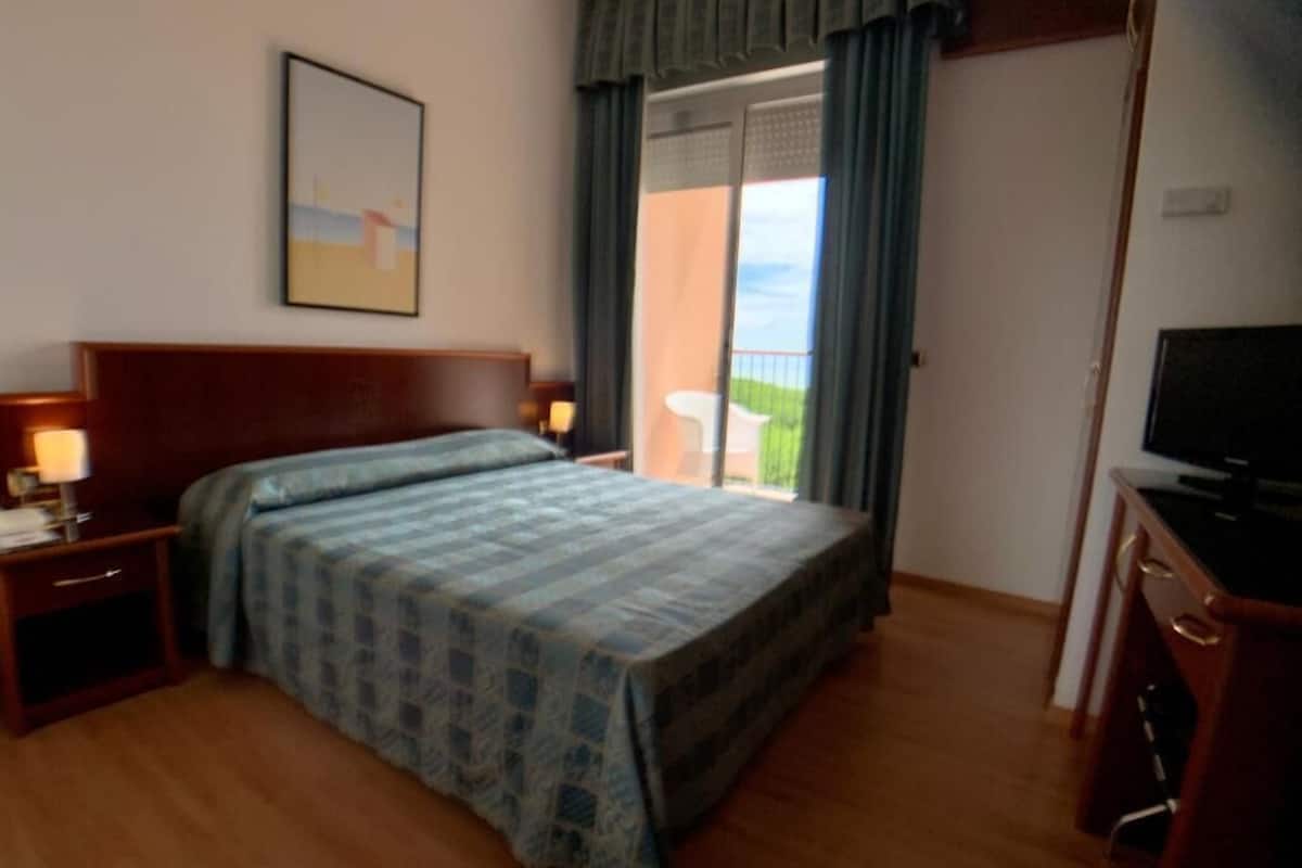 A bedroom with a bed, a nightstand, a dresser, a television, and a balcony with a view.