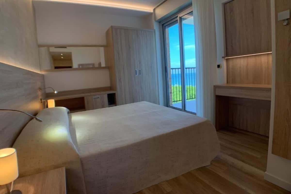 A modern bedroom with a large bed, a nightstand, a desk, and a wardrobe. There is a sliding glass door leading to a balcony with a view of the sky and greenery.