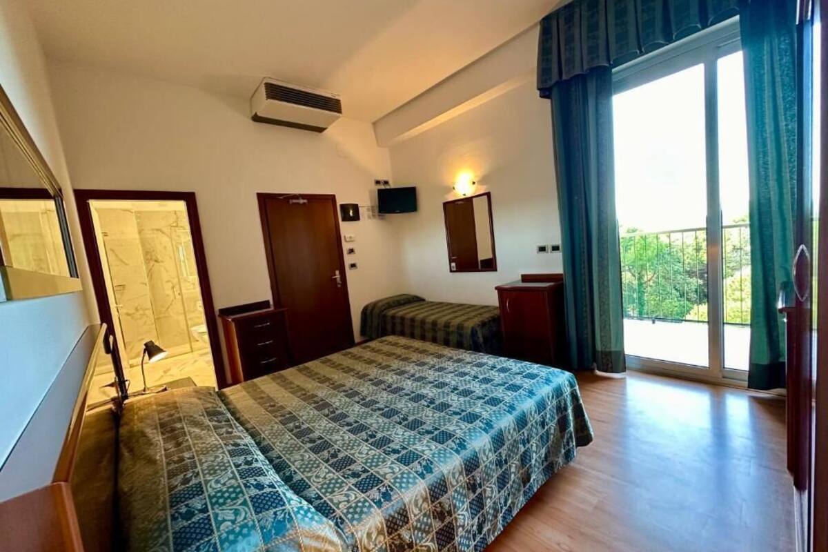 A bedroom with a large bed, a desk, a chair, a wardrobe, a TV, and a balcony with curtains.