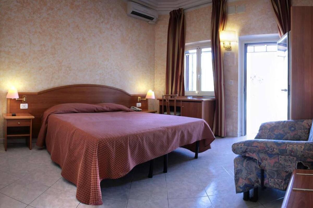 A hotel room with a large bed, two bedside lamps, a chair, a desk, and a window with curtains.
