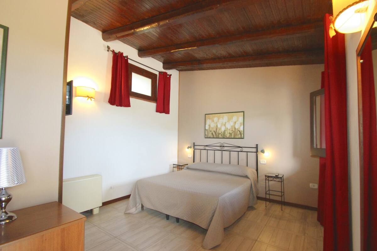 A bedroom with a bed, bedside lamps, a radiator, a wooden headboard, a painting on the wall, and a window with red curtains.