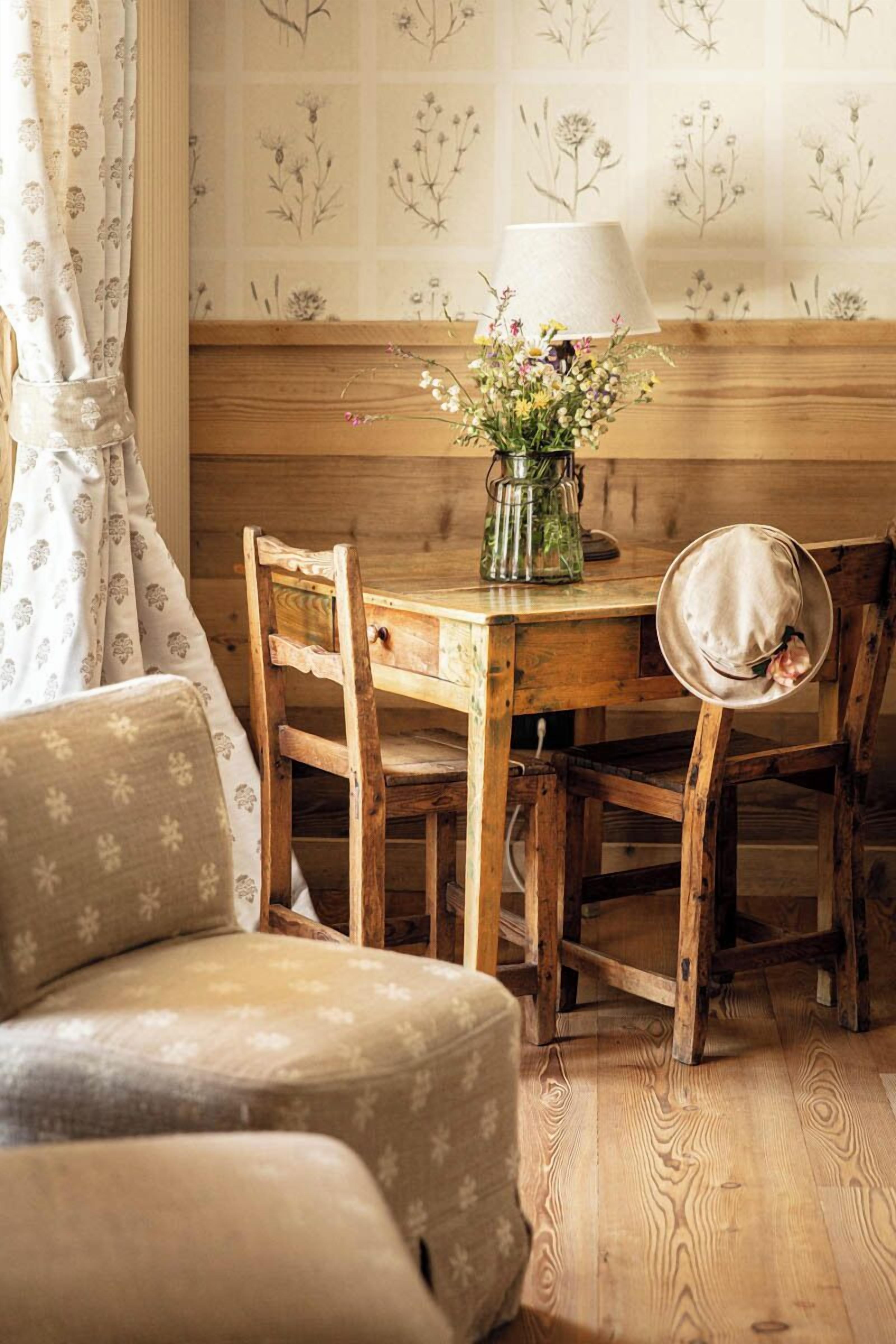 A wooden dining table with a vase of flowers and a lamp, set against floral wallpaper.