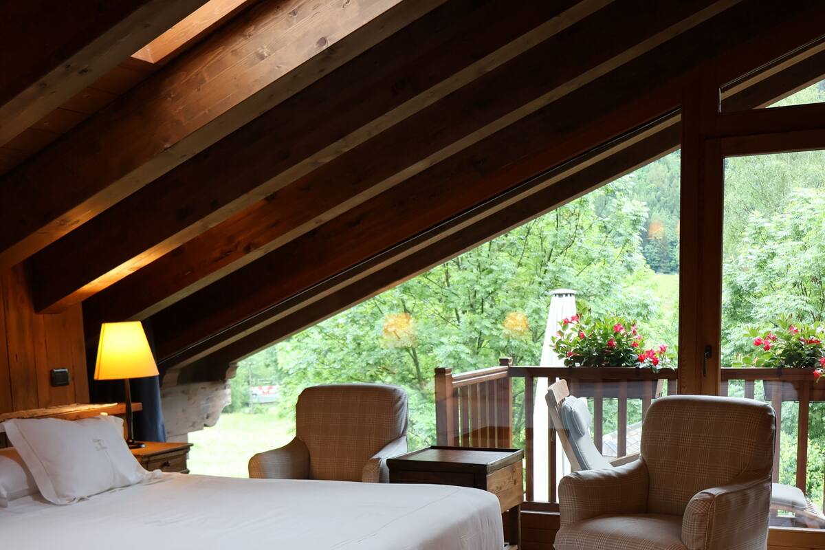 Un dormitorio con una cama grande, una silla, una mesita, una lámpara y vistas al exterior con vegetación.