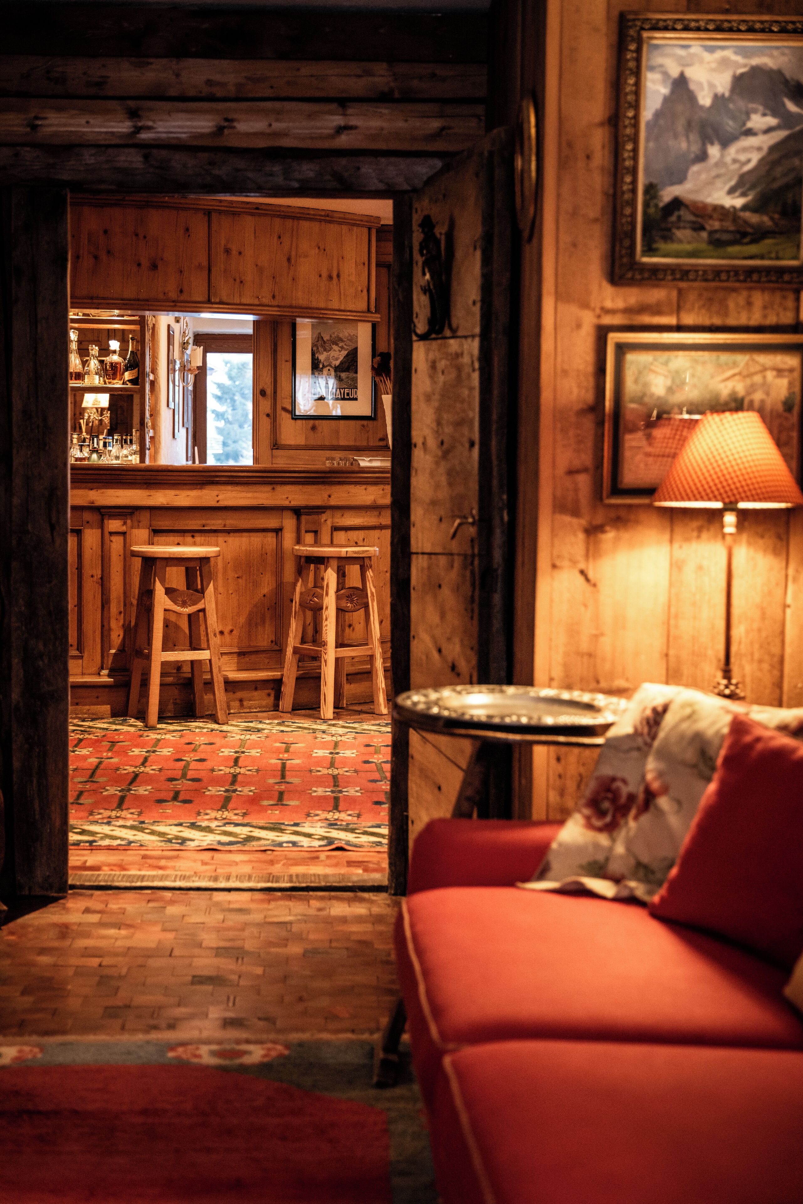 A cozy room with a wooden floor, a red sofa, a small table with a lamp, and a kitchen area with stools and a refrigerator.
