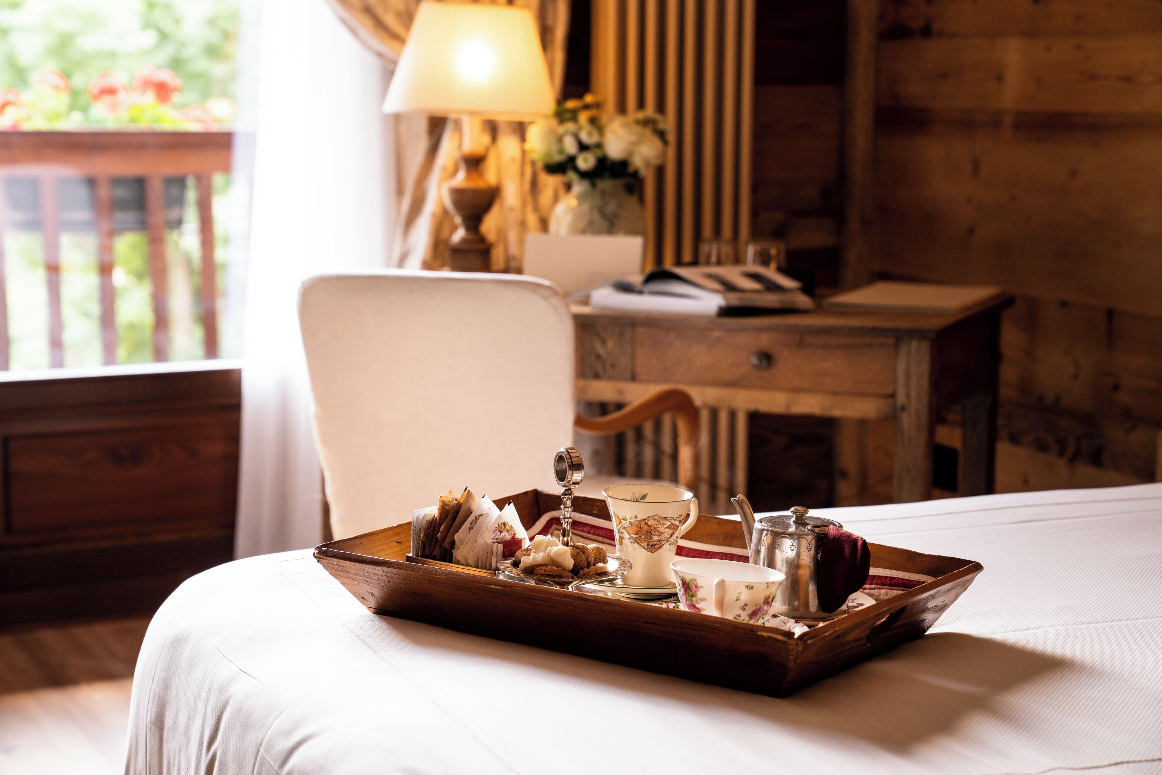 A neatly made bed with a tray of breakfast items, a lamp, and a desk with a chair in a room with wooden walls and a window.