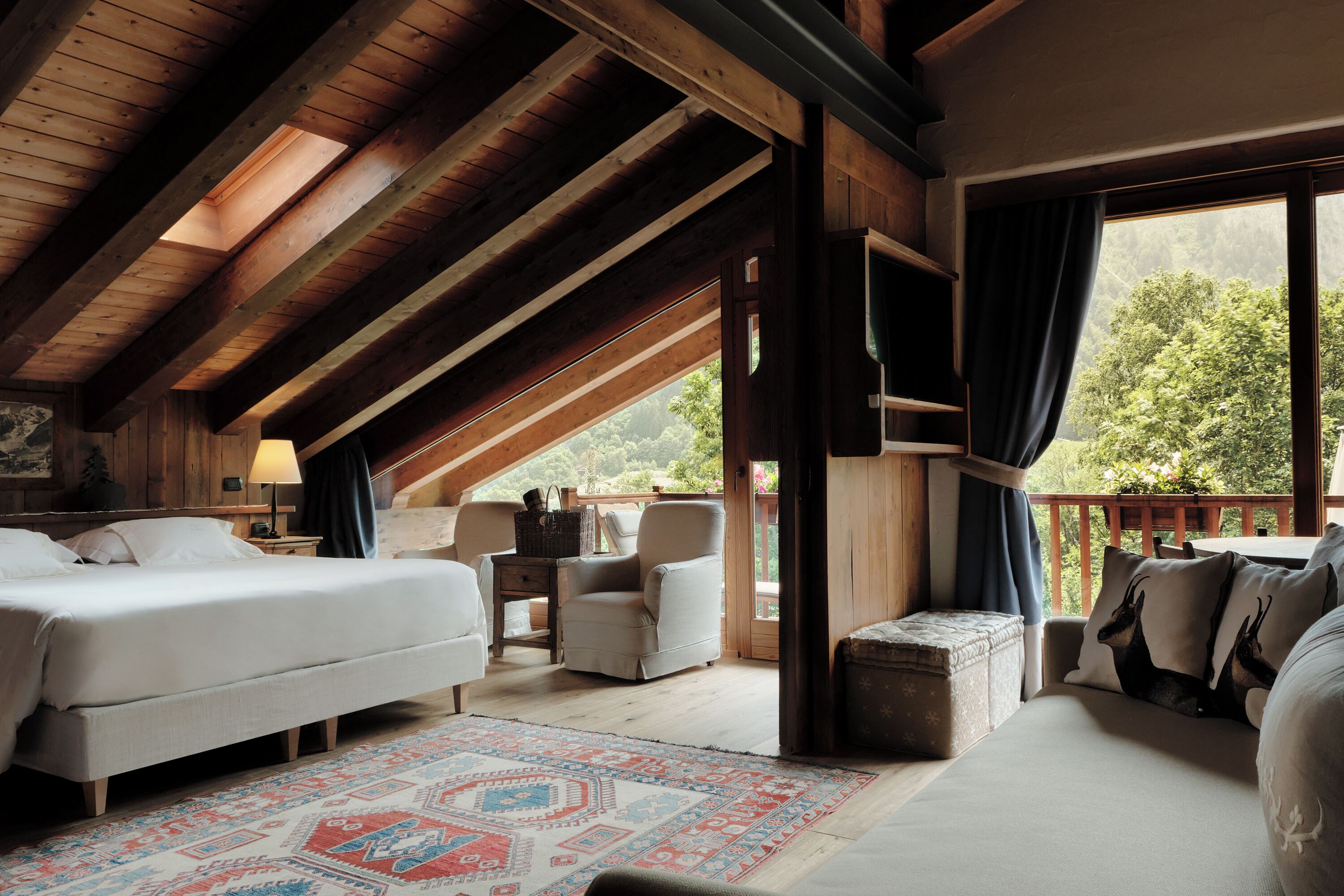 A spacious attic bedroom with a large bed, a seating area with a chair and table, and a balcony with a view of trees.