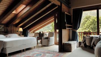 A spacious attic bedroom with a large bed, a seating area with a chair and table, and a balcony with a view of trees.