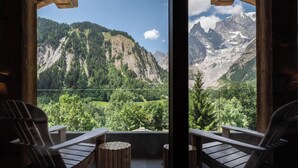 A wooden balcony with two chairs and a round wooden table, overlooking a mountainous landscape with green valleys and snow-capped peaks. - Auberge de La Maison (Courmayeur)