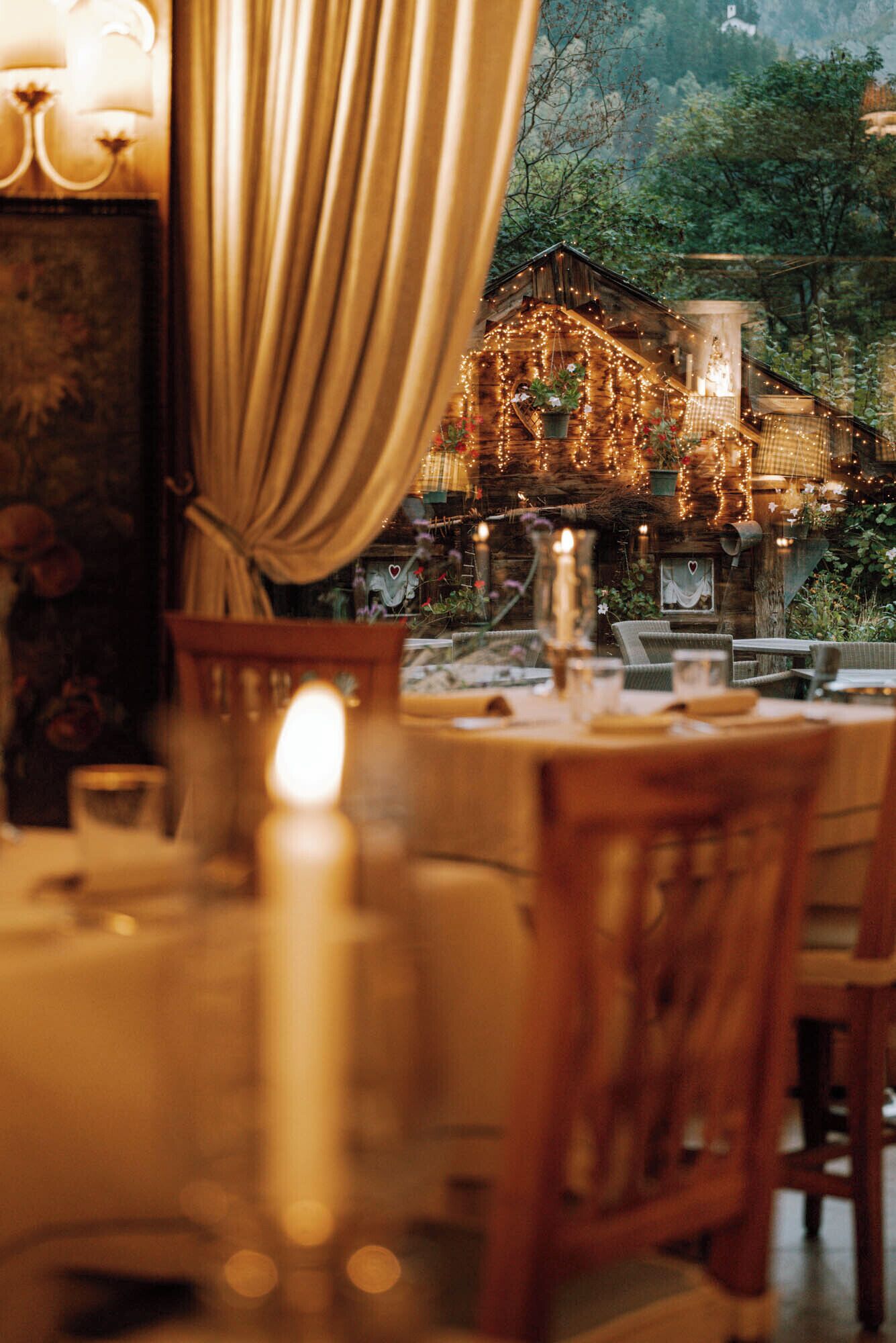 Un'accogliente zona pranzo all'aperto con tavoli e sedie in legno, un centrotavola con candela accesa e un edificio rustico sullo sfondo con luci decorative.