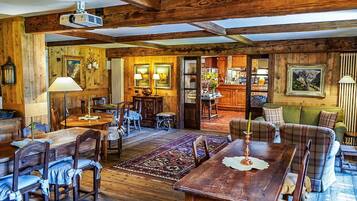 A rustic interior with wooden beams, a dining area with a large table and chairs, a bar area, and a cozy seating area with a patterned rug.