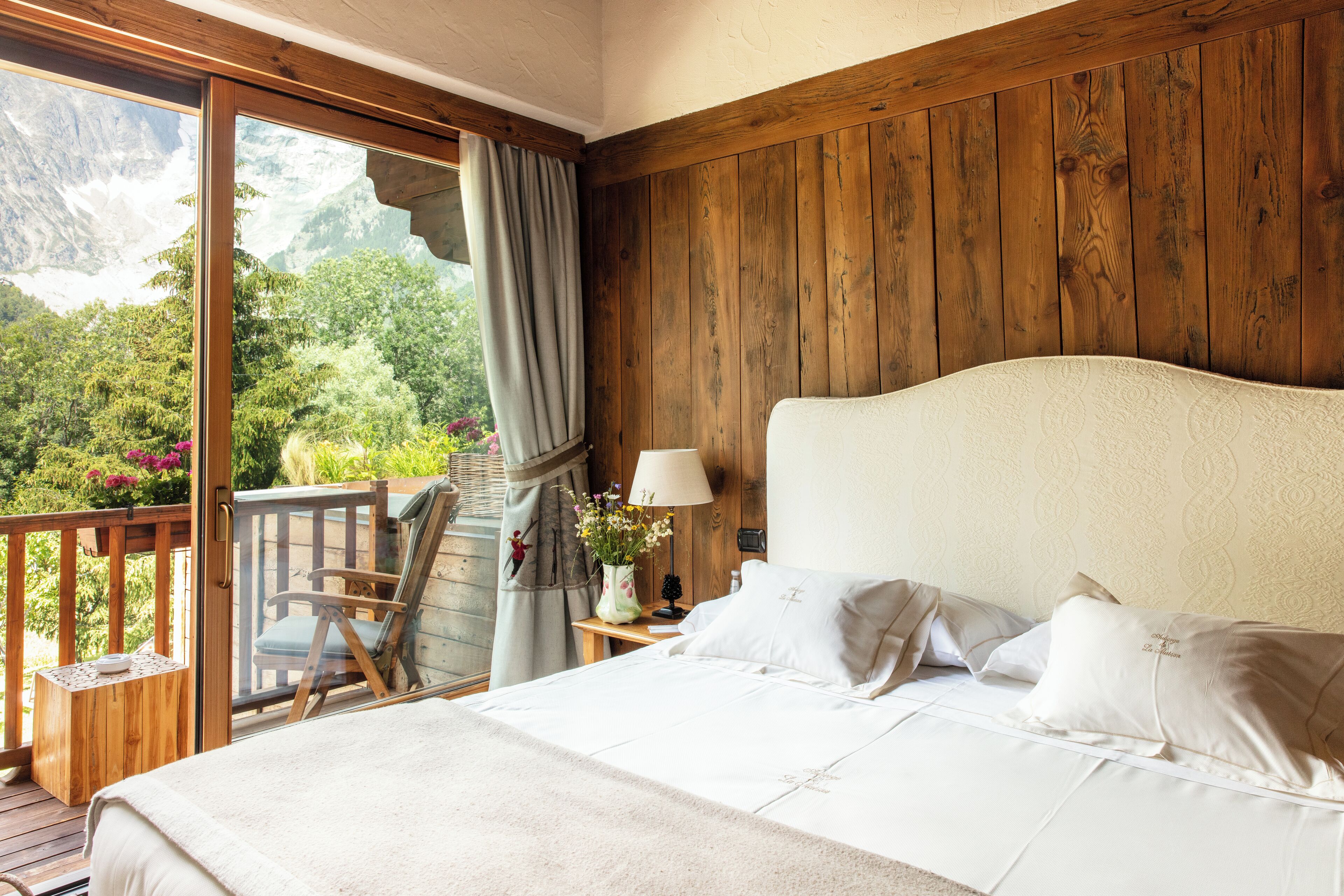 A bedroom with a large bed, a wooden headboard, a nightstand with a lamp, and a view of a balcony with a chair and a vase of flowers.
