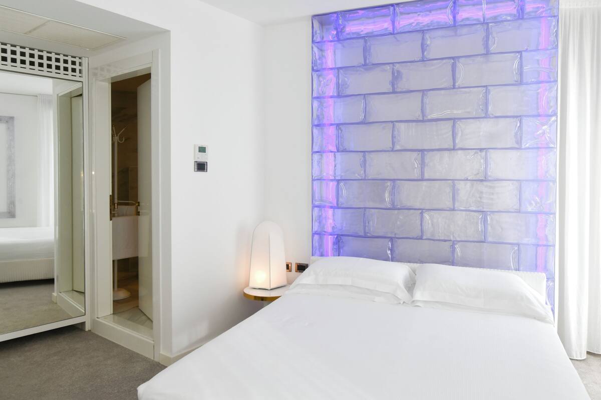 A hotel room with a bed, a nightstand with a lamp, a wall with a purple brick pattern, and a closet with a mirror.