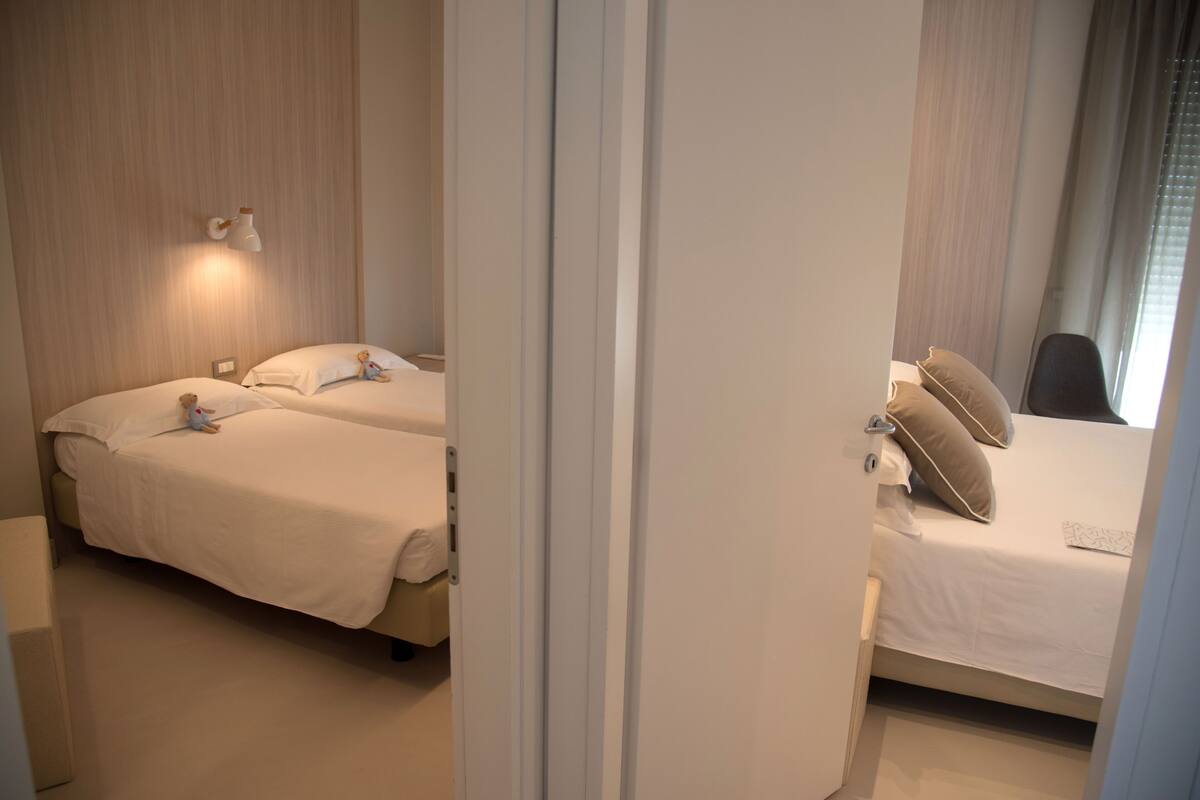 A hotel room with two beds, a nightstand, a lamp, and a window with curtains.