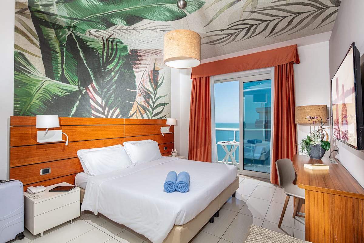 A hotel room with a bed, a desk, a chair, a small table, a nightstand, a lamp, and a view of the balcony.