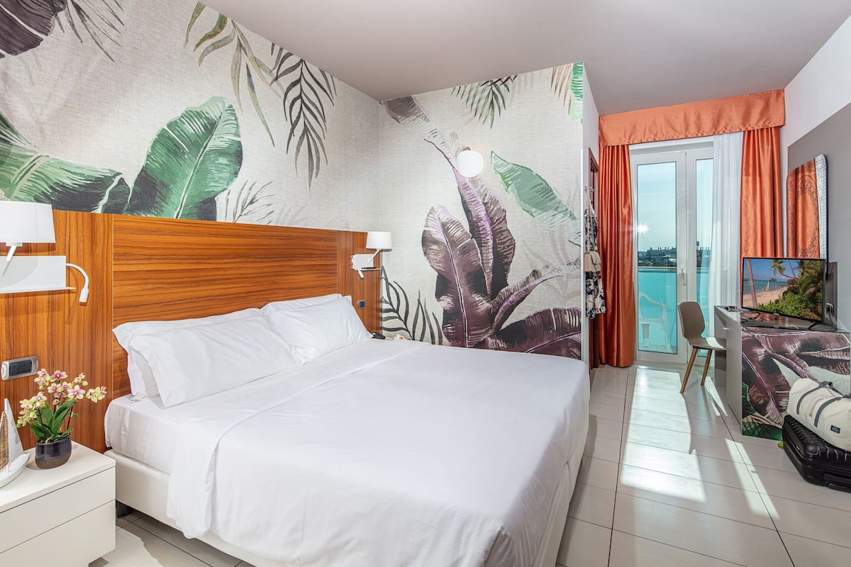 A hotel room with a large bed, a wooden headboard, a wall mural with tropical leaves, a balcony with orange curtains, and a television on a desk.