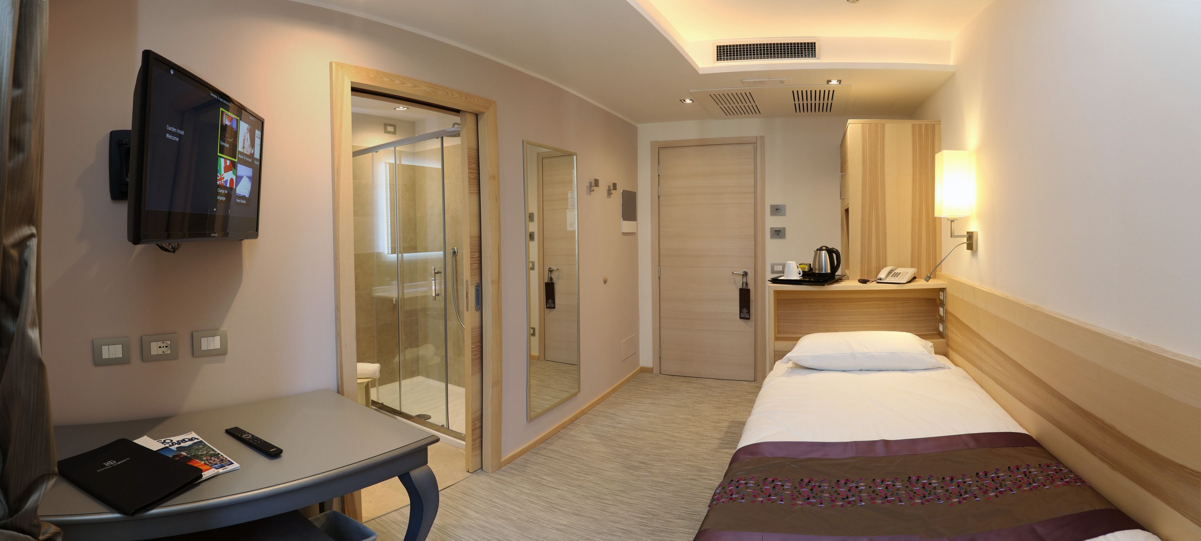 A hotel room with a bed, a desk with a remote control, a television, a mirror, a wardrobe, and a bathroom with a glass door.