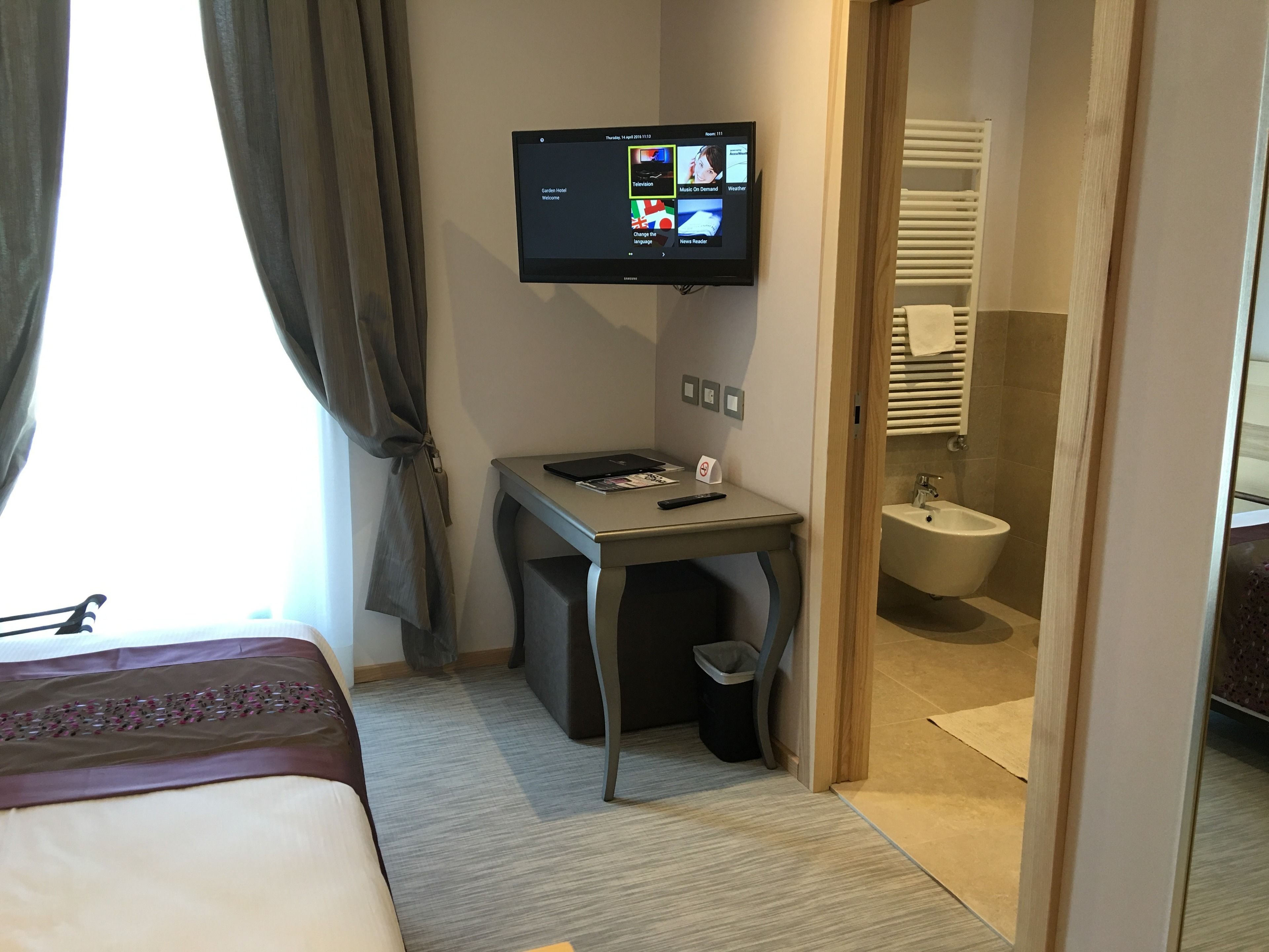 A hotel room with a bed, a desk with a computer, a TV mounted on the wall, and a bathroom visible through an open door.