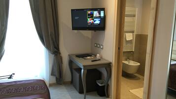 Ein Hotelzimmer mit einem Bett, einem Schreibtisch mit Computer, einem an der Wand befestigten Fernseher und einem durchgegangenen Tür erkennbaren Badezimmer.