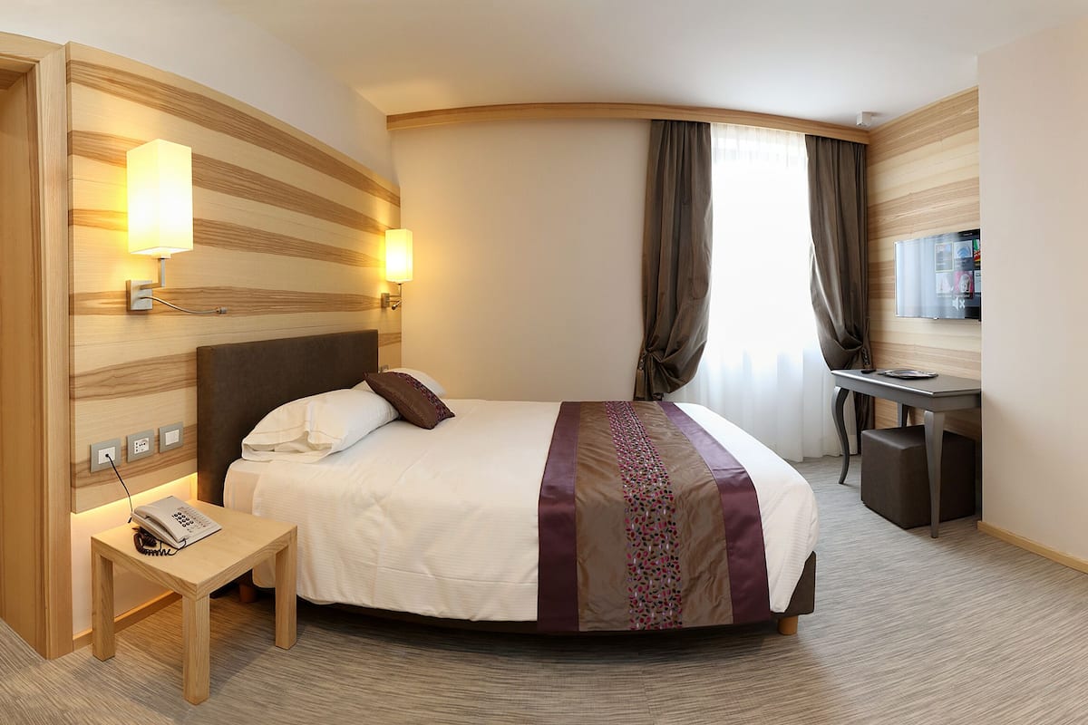 A hotel room with a bed, bedside table, lamp, and a desk with a chair.