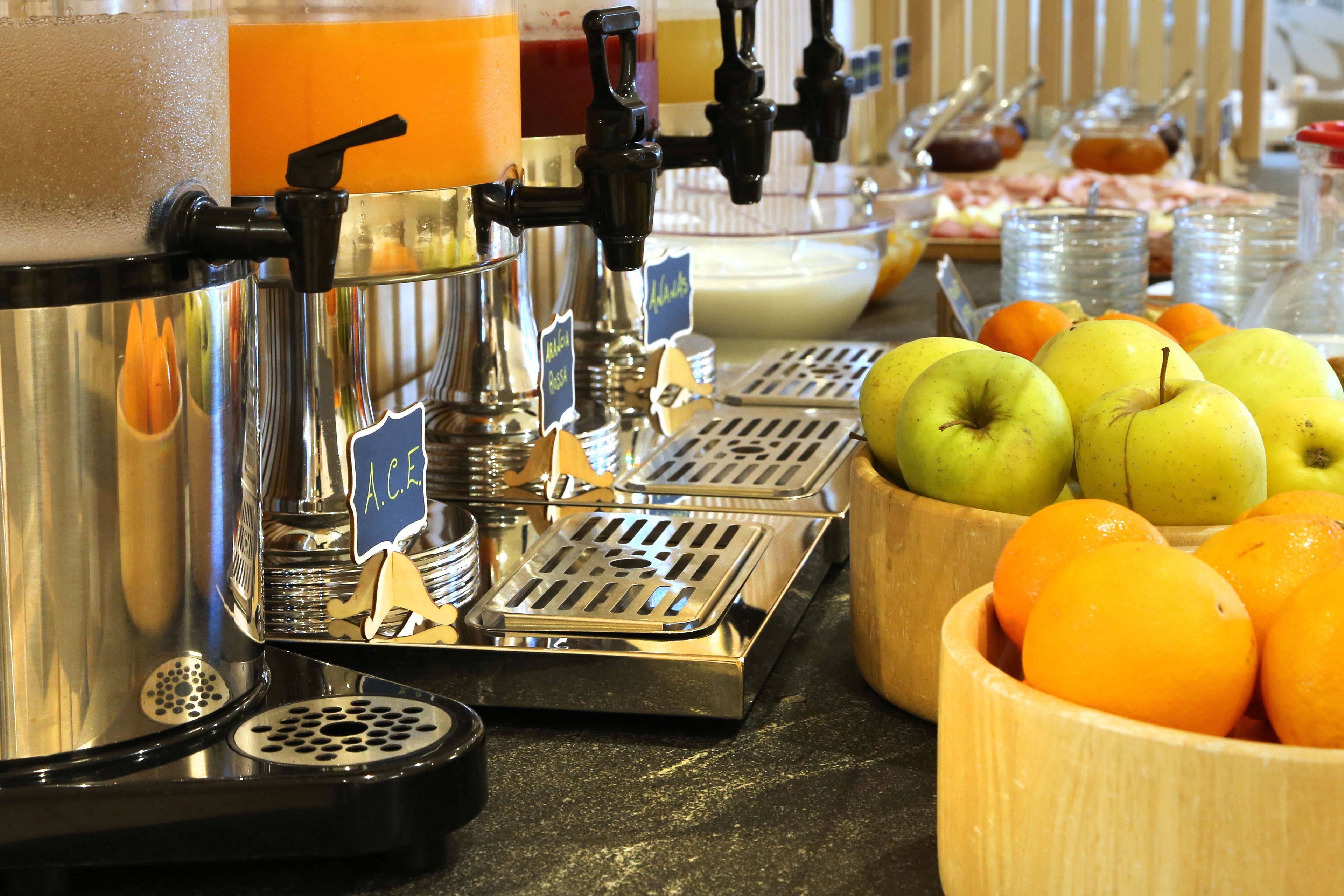A coffee machine with multiple dispensers, a bowl of apples, and a bowl of oranges on a counter.