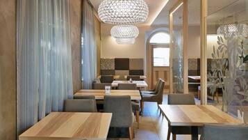 Un comedor moderno con mesas de madera y sillas grises tapizadas, una gran lámpara de cristal y vistas a la cocina a través de un separador de vidrio.