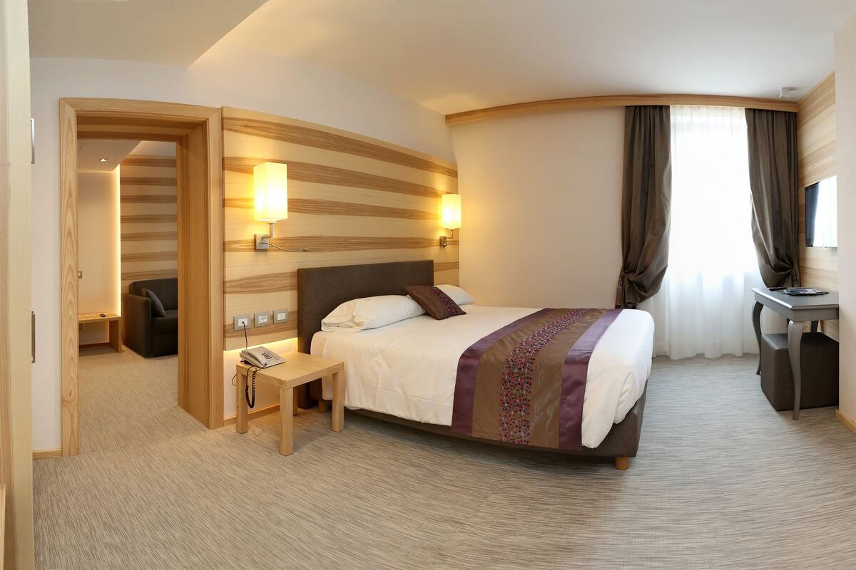 A hotel room with a bed, bedside table, lamp, desk, and a shower area.