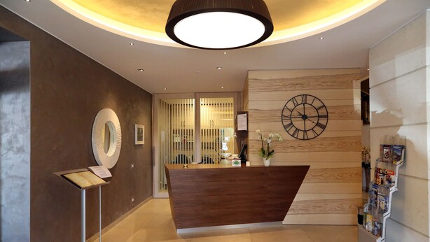 A modern reception area with a wooden reception desk, a large clock, and a magazine rack. - Hotel Garden (Peschiera del Garda)