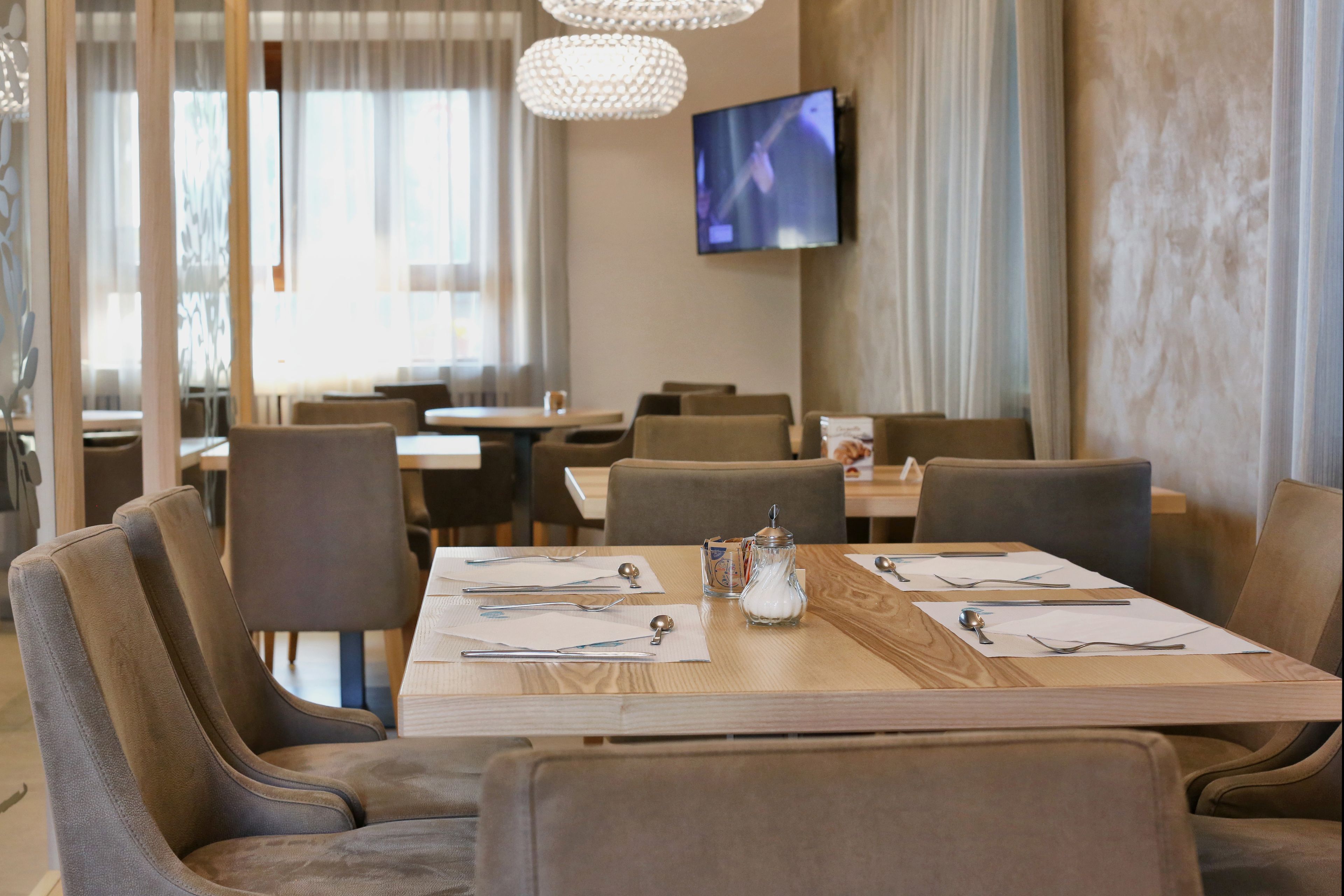 A dining area with a long wooden table set for four, surrounded by upholstered chairs. A television is mounted on the wall, and there is a window with sheer curtains.