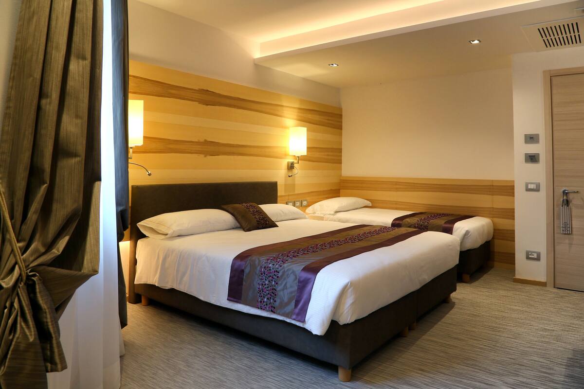 A hotel room with a large bed, two bedside tables, a headboard, a wall-mounted lamp, and a door.