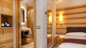 A hotel room with a wooden wardrobe, a bathroom with a sink and mirror, a bedside table with a lamp, and a bed with a headboard. - Hotel Garden (Peschiera del Garda)