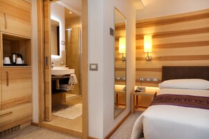 Habitación de hotel con armario de madera, baño con lavabo y espejo, mesita de noche con lámpara y cama con cabecero.