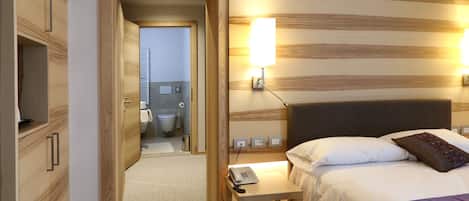 A hotel room with a wooden headboard, a bedside table with a lamp, a bed with white bedding, and a view into the bathroom.