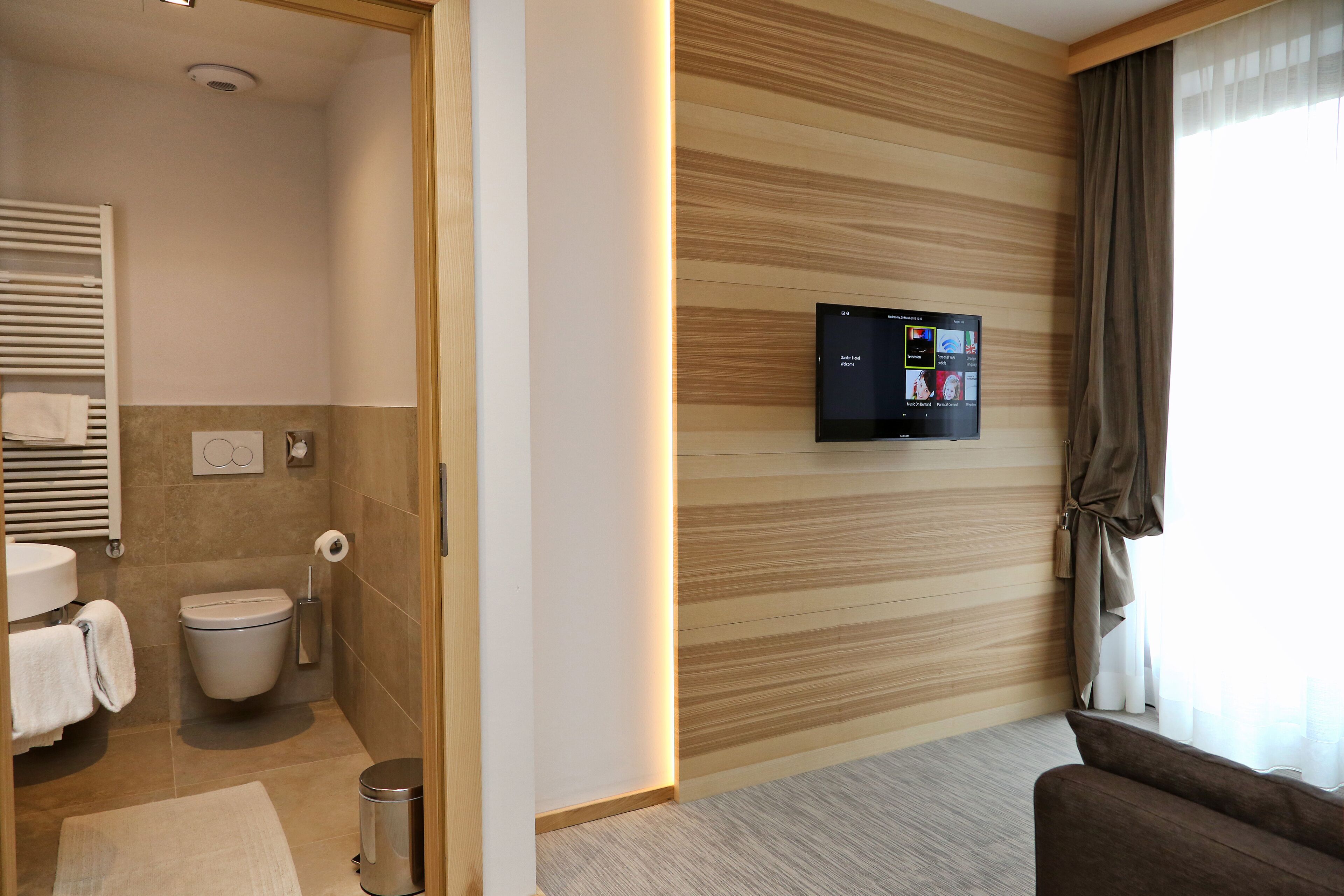 A modern bathroom with a toilet, sink, and towel rack. A flat-screen TV is mounted on the wall, and a window with curtains is visible.