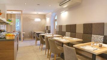 A modern dining area with wooden tables and chairs, a kitchen counter with fruits, and a patterned wall.