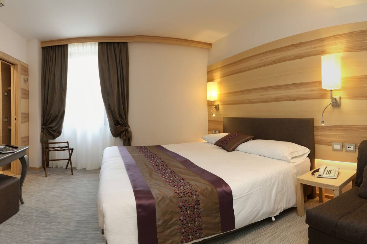 A hotel room with a bed, a desk, a chair, a TV, and a nightstand with a lamp.