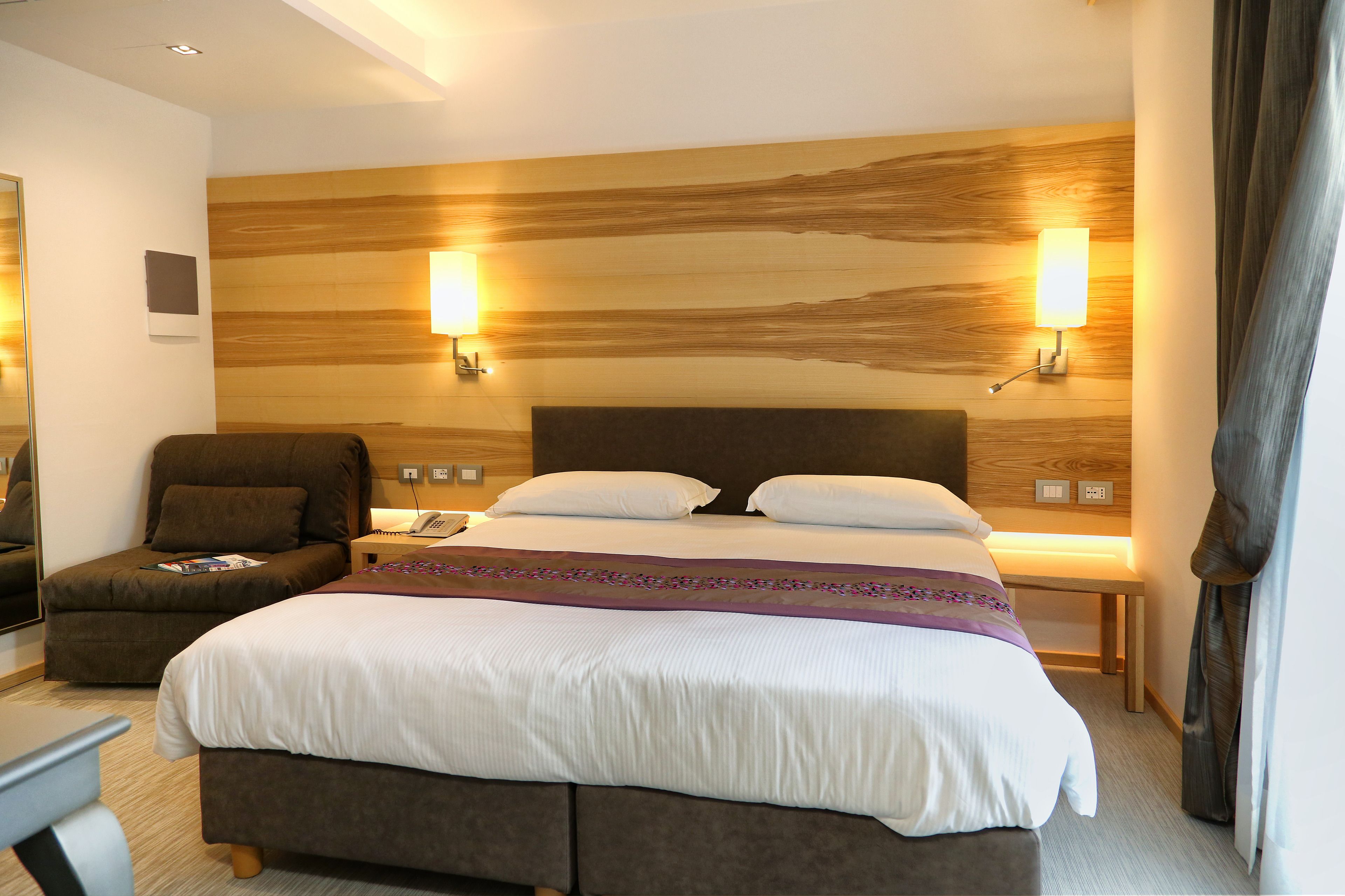 Una habitación de hotel con una cama grande, dos lámparas de noche, una mesita de noche, una silla y un cabecero de madera.