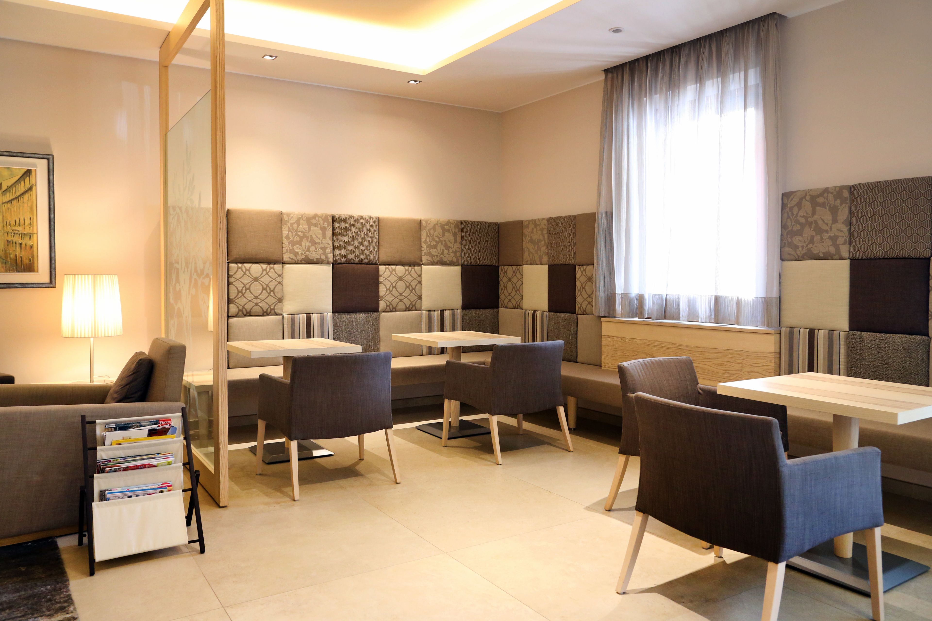 A modern hotel room with a seating area featuring cushioned benches and tables, a bookshelf, and a large window with curtains.