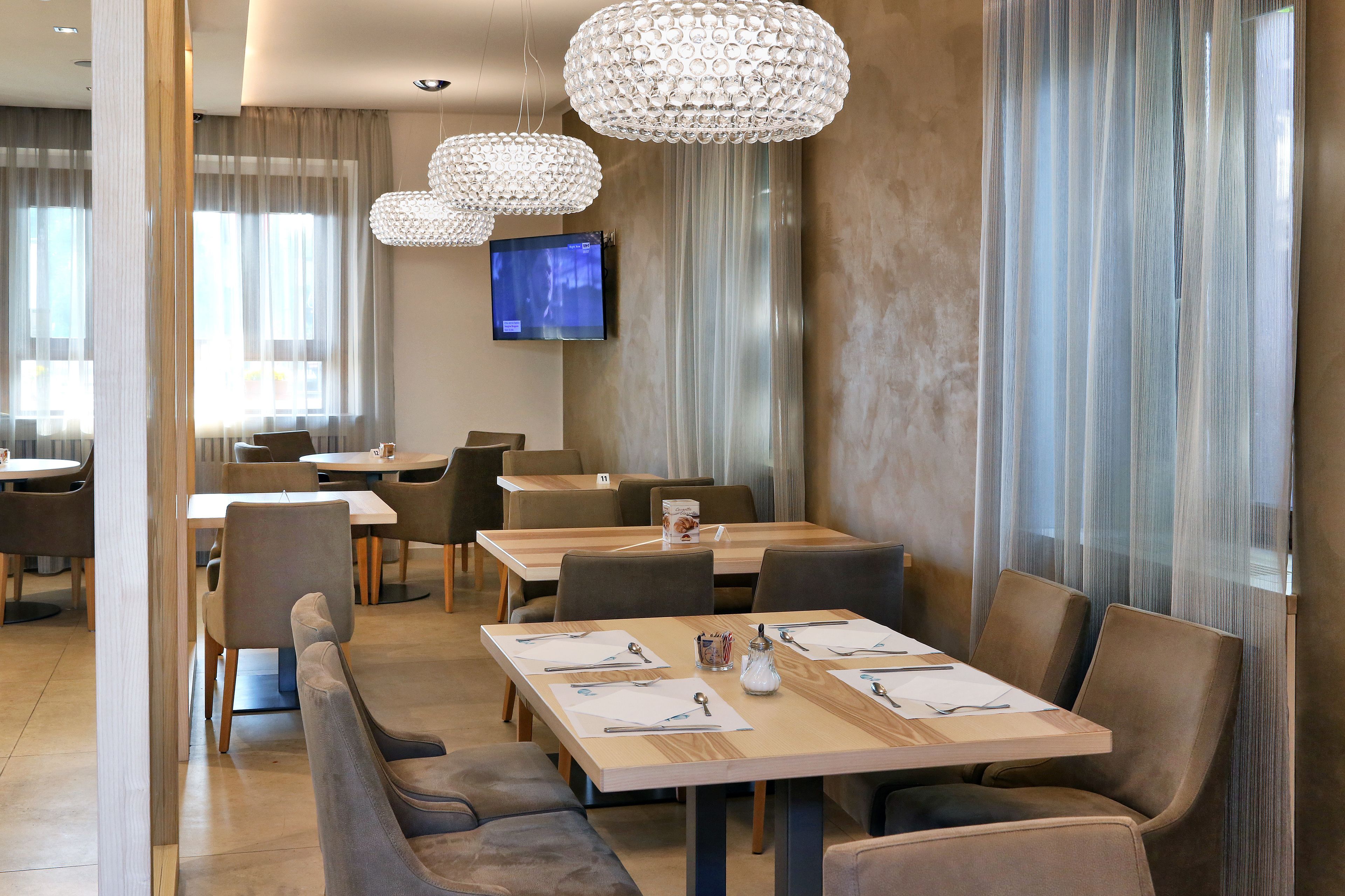 A modern dining area with multiple tables and chairs, a television mounted on the wall, and decorative crystal chandeliers.