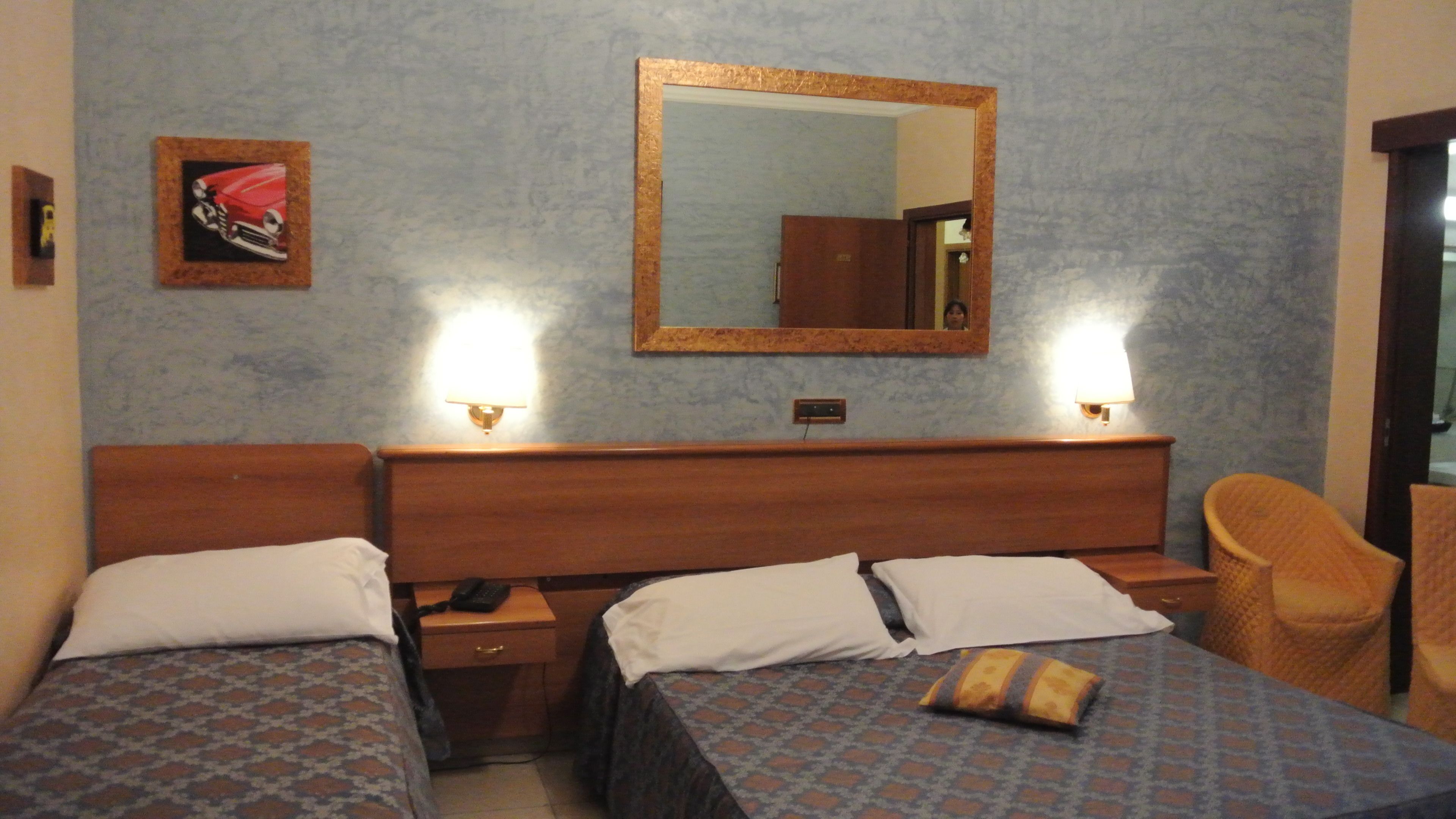 Une chambre d’hôtel avec deux lits, une tête de lit en bois, un miroir et une lampe fixée au mur.