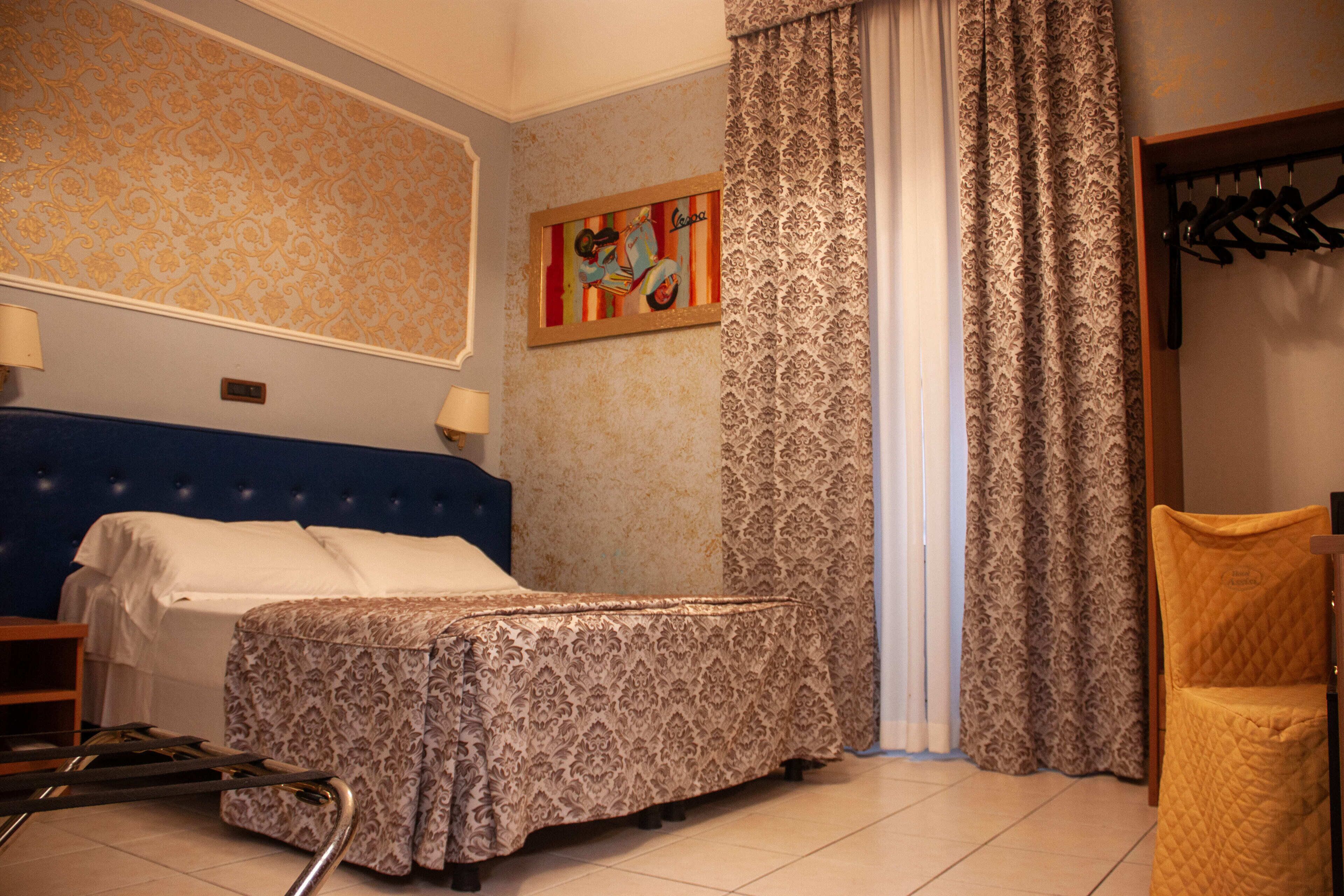 Una habitación de hotel con cama, una silla, perchero y un cuadro en la pared.