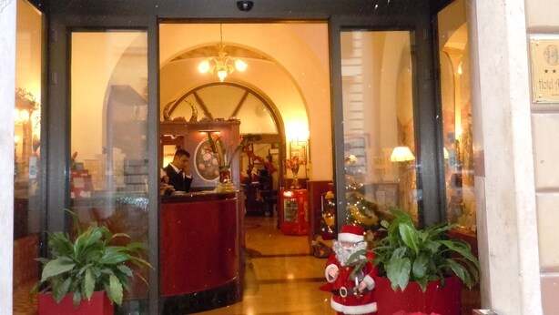 The entrance of Hotel Assisi with a reception desk, a chandelier, and potted plants. - Hotel Assisi (Rome)