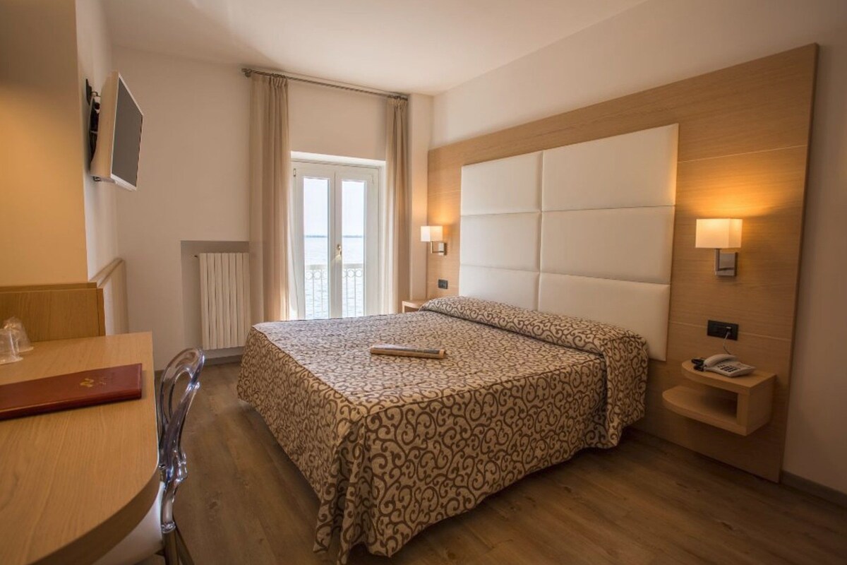 Habitació d'hotel amb un llit, una taula de nit, una làmpada i vista a l'exterior.