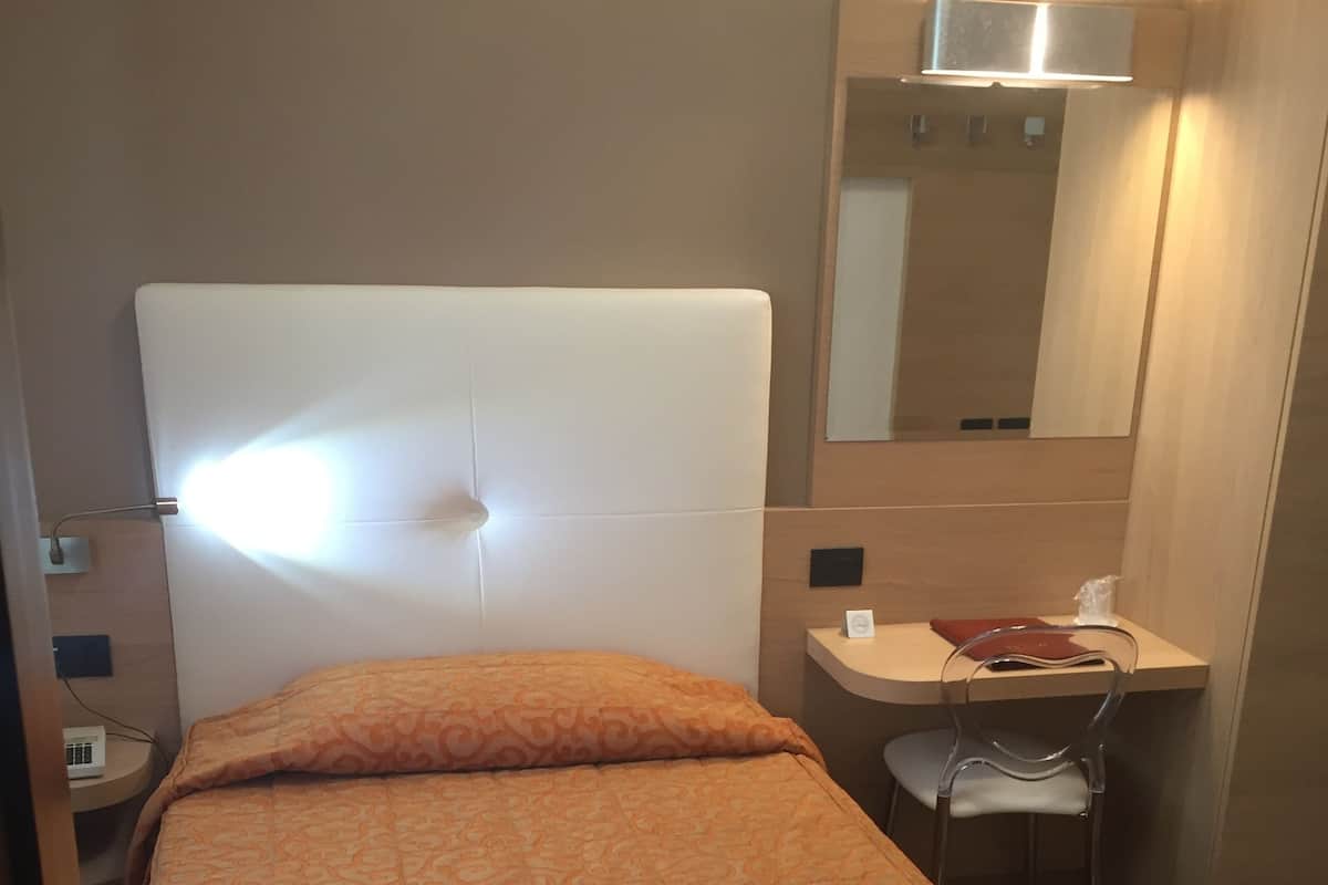 Een eenpersoonsbed met een oranje bedspreide, een nachtkastje met een lamp, een spiegel boven de spiegel en een klein bureau met een stoel.
